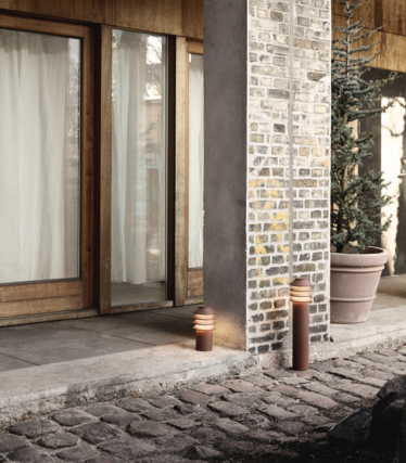 Ulkona oleva kohtaus, jossa kaksi modernia sylinterimäistä polkuvaloa on sijoitettu kivisen kävelytien ja tiili- ja betonipylvään viereen. Niiden takana on puurunkoiset lasiovet, joissa on valoverhot, sekä suuri ikivihreä ruukkukasvi. Valojen lämmin hehku luo kodikkaan ja kutsuvan tunnelman.